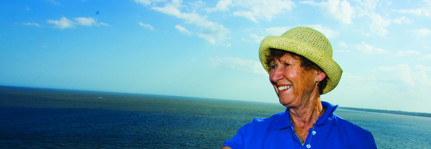 A smiling woman in a straw hat stands on a historic lighthouse balcony overlooking the coast of St. Simons Island, Georgia.