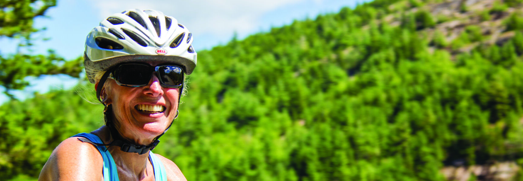 A smiling woman wearing a bicycle helmet and sunglasses enjoys a bicycling adventure against a backdrop of lush green mountains in Acadia National Park, Maine.