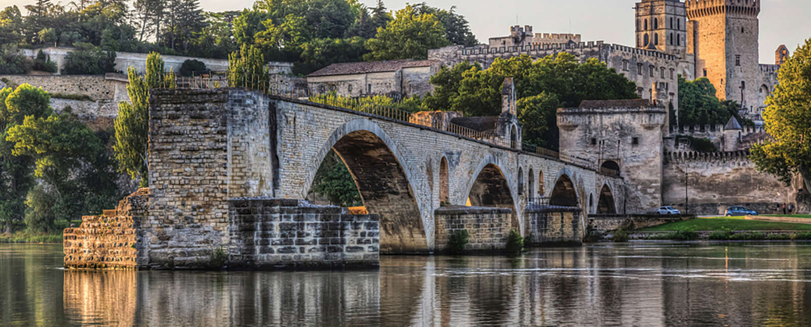 A historic stone bridge with several arches crosses the Rhône River in France, with a grand medieval palace and cathedral visible in the background.