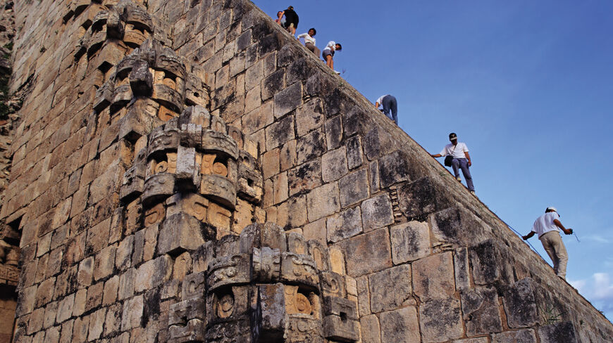 Tourists climb the steep stone steps of an ancient Maya ruin in Mexico's Northern Yucatan, featuring detailed stone-carved masks along the side.