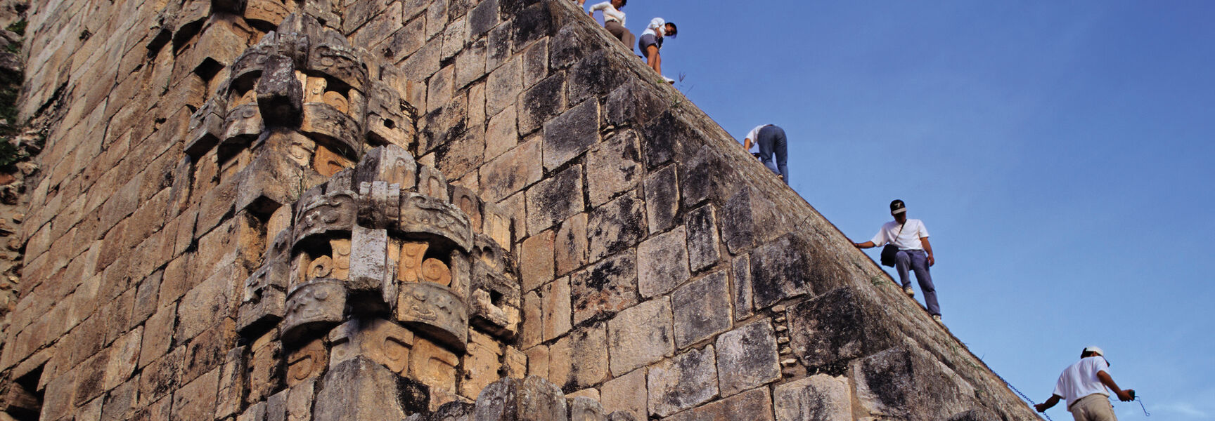 Tourists climb the steep stone steps of an ancient Maya ruin in Mexico's Northern Yucatan, featuring detailed stone-carved masks along the side.