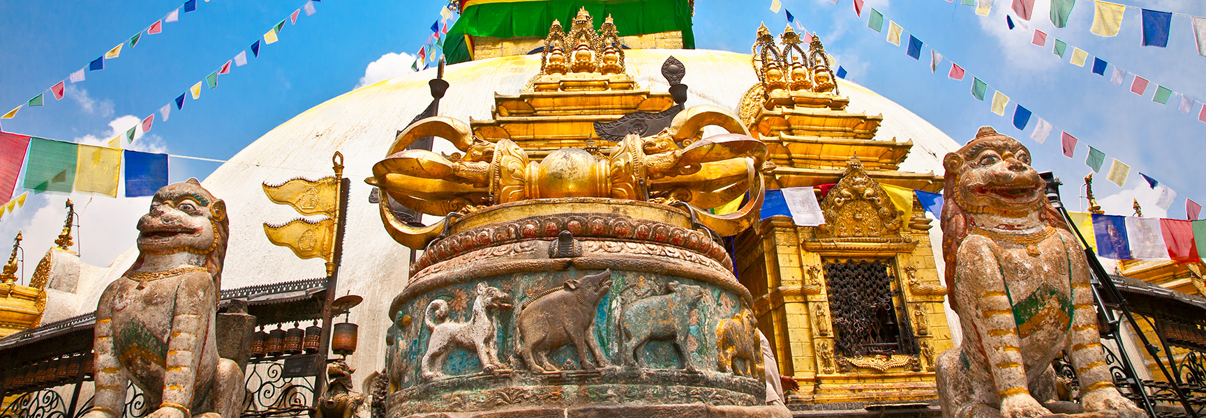 Stone lion statues guard the ornate golden Swayambhunath temple in Nepal, with colorful prayer flags flying against a blue sky.