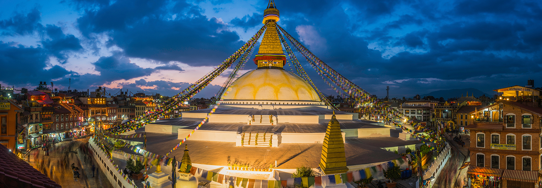 The Boudhanath Temple stupa in Nepal glows at dusk, with prayer flags radiating from its golden spire over the surrounding city of Kathmandu.