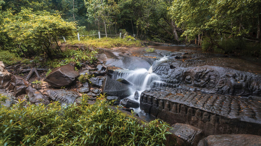 Water cascades over ancient stone carvings of Khmer deities in a forested riverbed near the temples of Angkor in Cambodia.