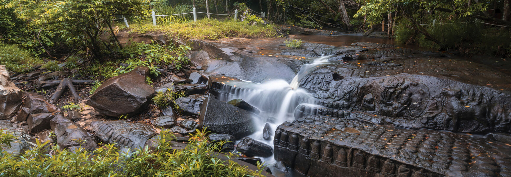 Water cascades over ancient stone carvings of Khmer deities in a forested riverbed near the temples of Angkor in Cambodia.
