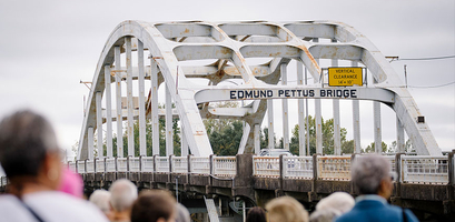 A crowd of people looks toward the large, white-arched Edmund Pettus Bridge in Selma, Alabama on an overcast day.