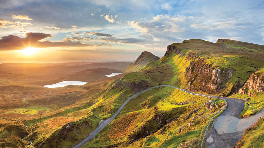 A winding road snakes through the vibrant green mountains of the Scottish Highlands during a scenic sunset overlooking distant lakes.