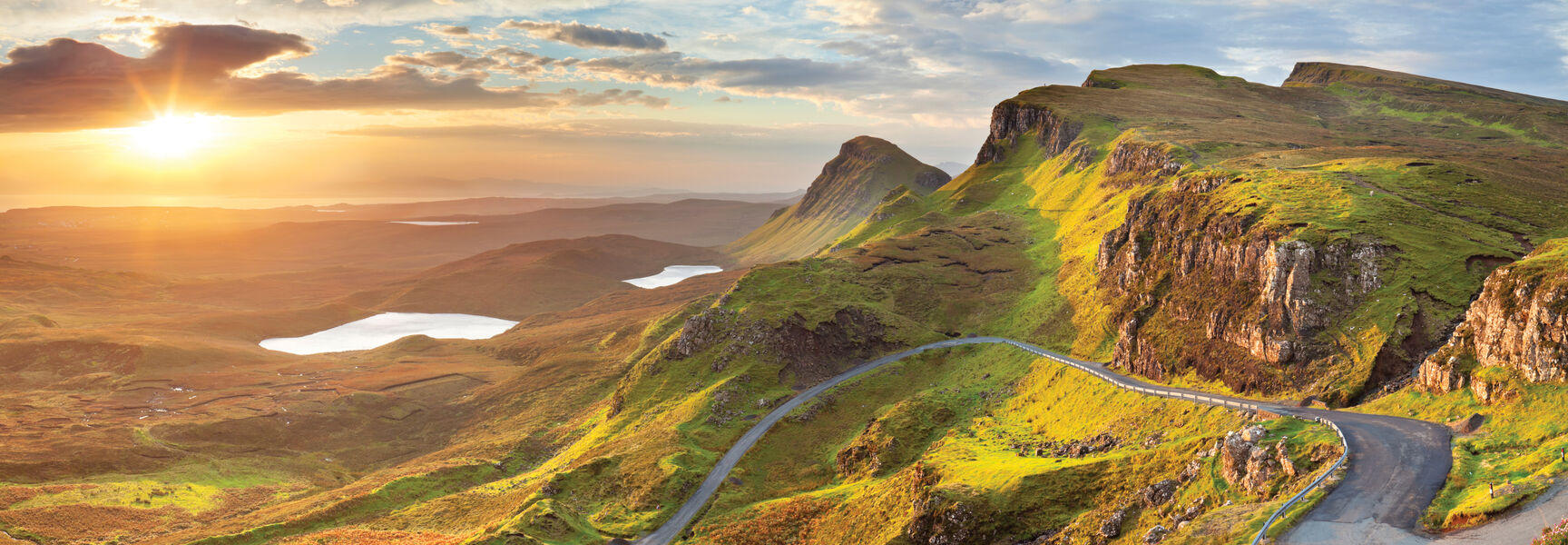 A winding road snakes through the vibrant green mountains of the Scottish Highlands during a scenic sunset overlooking distant lakes.