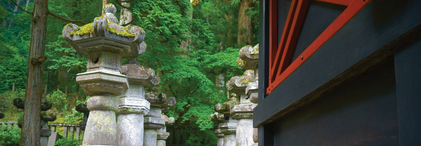 Traditional stone lanterns and an elaborate red and black shrine stand in a lush, green forest in Northern Japan.