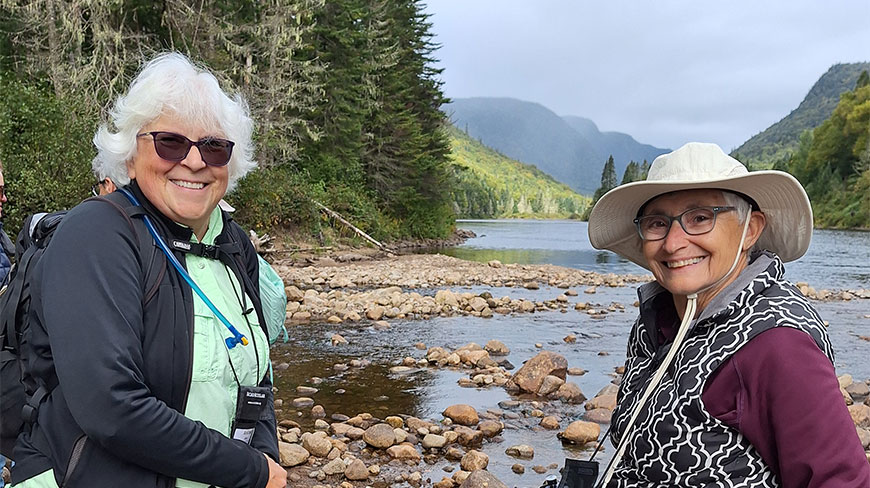 Two women in hiking gear smile while taking a break by a rocky river in a scenic Québec park.
