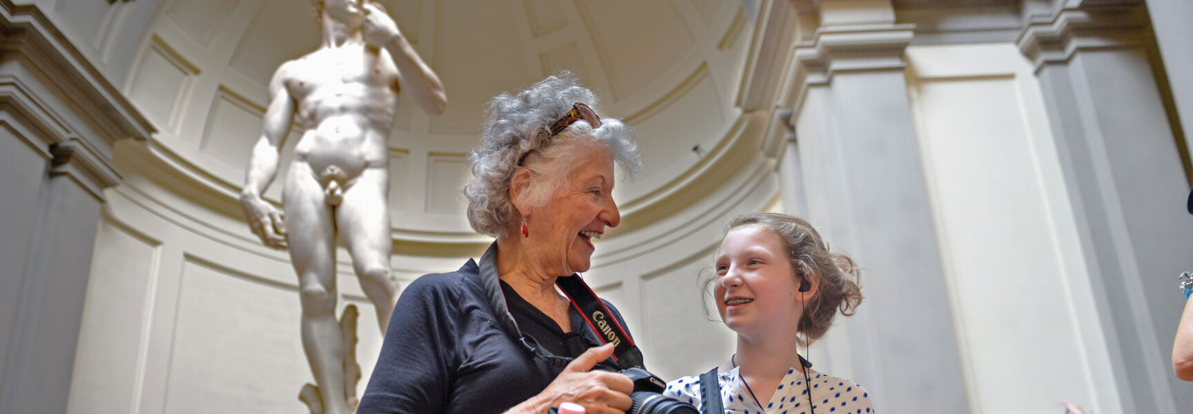 An older woman and young girl smile together in front of Michelangelo's David statue at the Accademia Gallery in Florence, Italy.