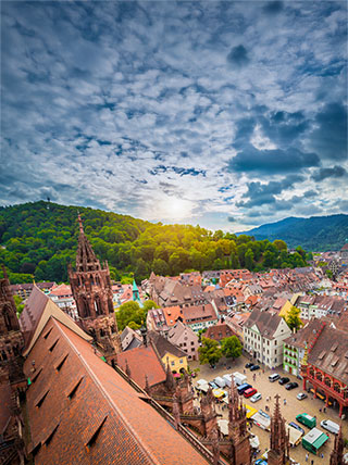 High-angle view of the historic town square in Freiburg, Germany, with the cathedral and forested hills under a dramatic, cloudy sky.
