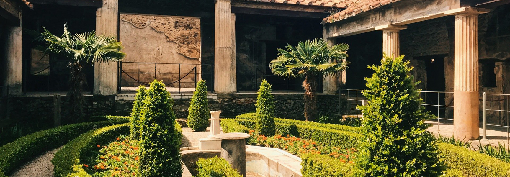 A lush courtyard garden with manicured hedges and palm trees surrounded by the ancient stone ruins of a villa in Pompeii, Italy.