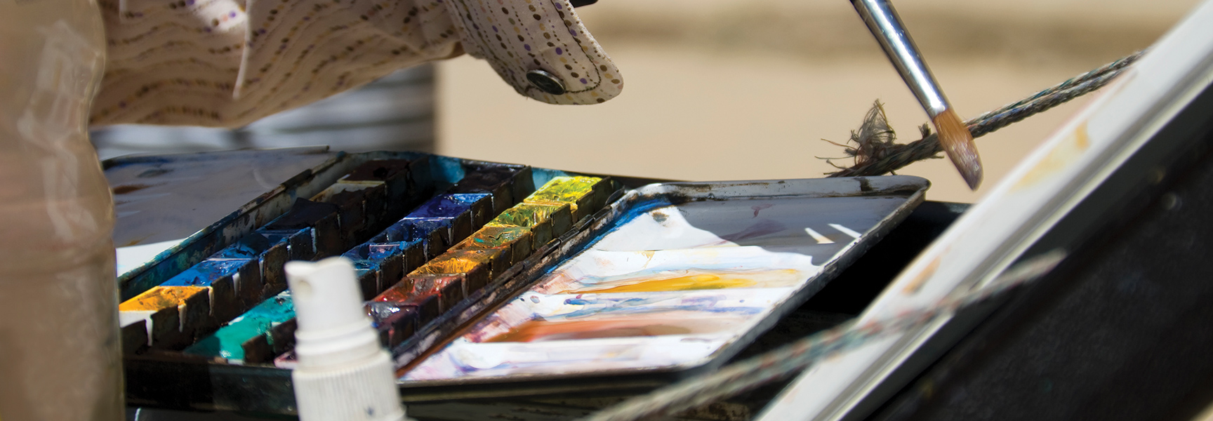 An artist holds a paintbrush over a well-used watercolor palette while painting outdoors in California.