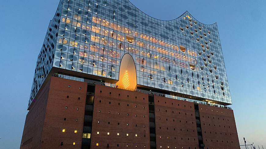 The modern glass facade of the Elbphilharmonie concert hall in Hamburg, Germany, glows at dusk above its historic brick base.