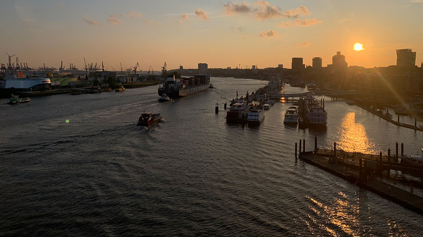 Boats and a large container ship navigate the Hamburg Harbour in Germany at sunset, with the city skyline silhouetted against the golden sky.