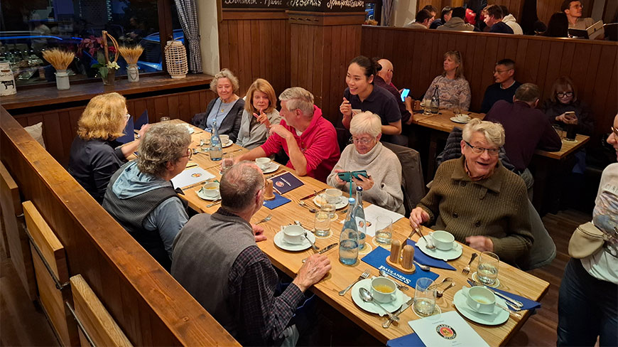 A tour group enjoys a meal together at a long wooden table inside a cozy, traditional-style restaurant in Germany.