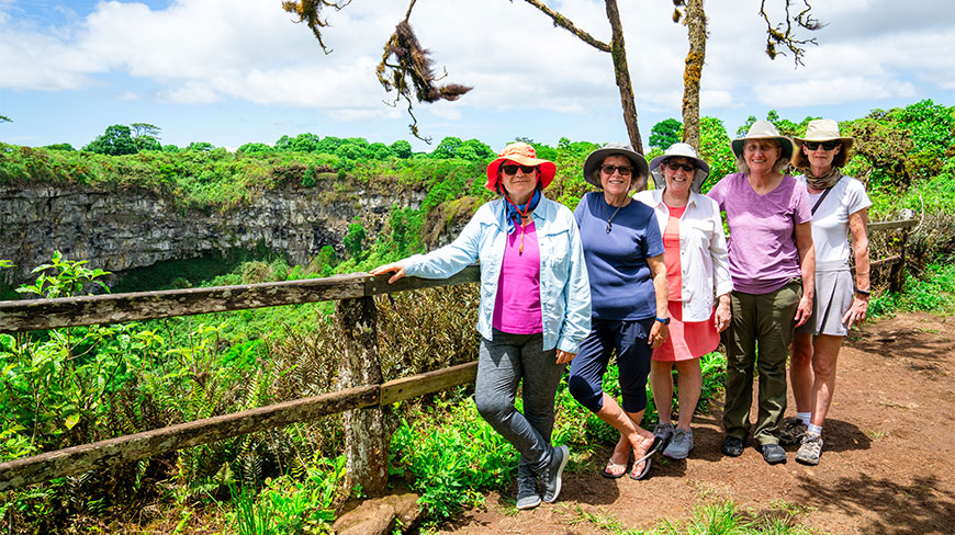 Five women wearing sun hats stand at a wooden railing overlooking a lush green crater in the Galapagos Santa Cruz Highlands.