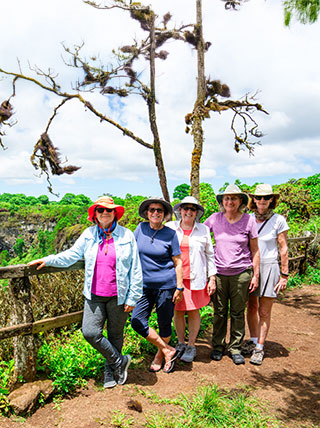 Five women wearing sun hats stand at a wooden railing overlooking a lush green crater in the Galapagos Santa Cruz Highlands.