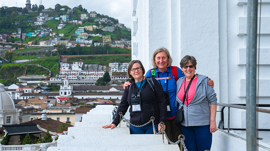 Three women on a Quito walking tour smile together at the San Francisco Church and Convent with the city and hillside in the background.