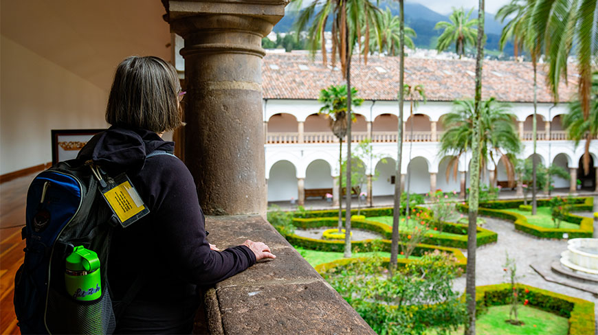 A traveler with a backpack overlooks the manicured gardens and arched corridors of the San Francisco Church and Convent during a Quito walking tour.