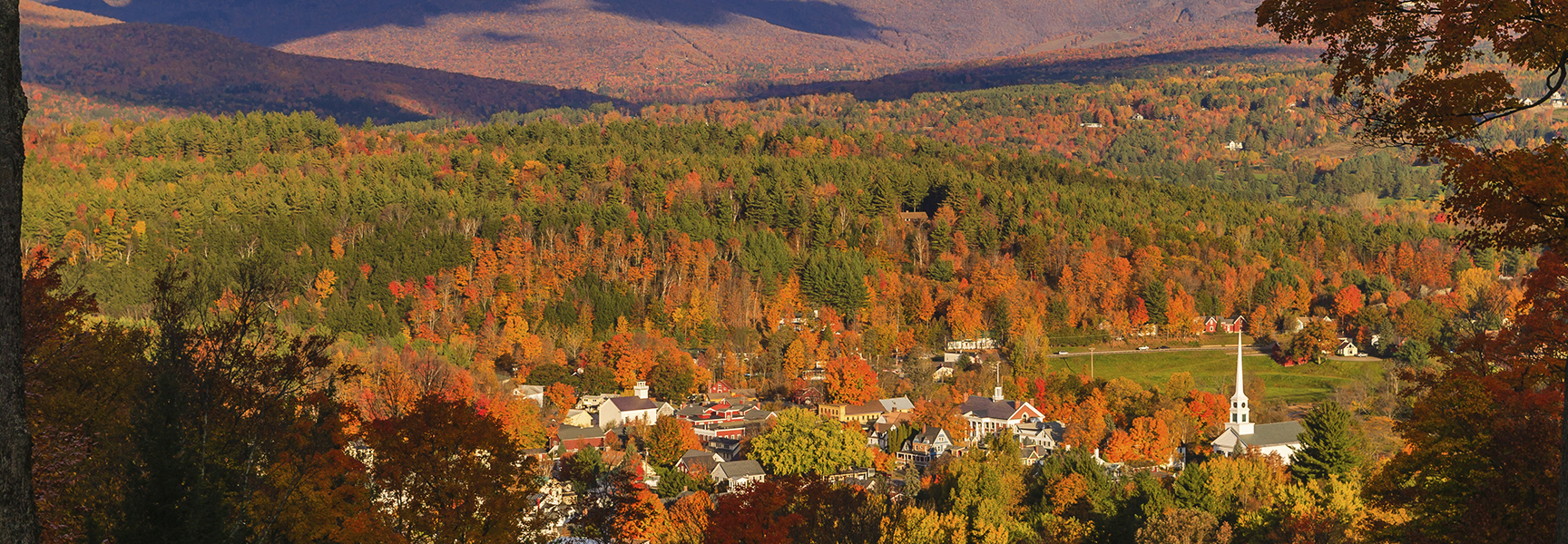 A small town with a church steeple is nestled in a valley of vibrant autumn foliage in Stowe, Vermont.