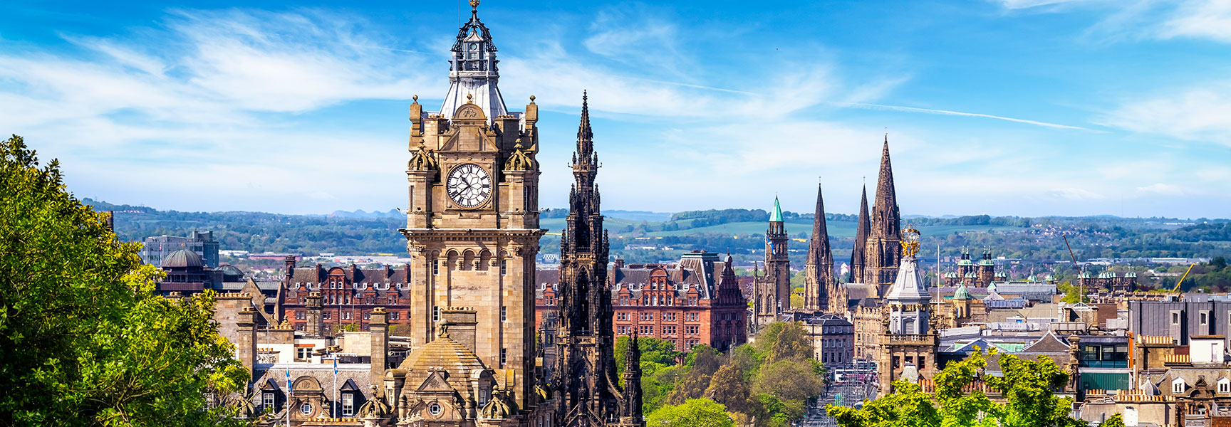 A scenic view of the historic Edinburgh, Scotland skyline, featuring the Balmoral clock tower and other spires under a bright blue sky.