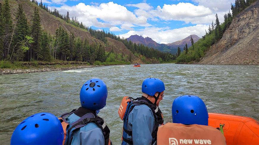 Several rafters in blue helmets float down a river in Alaska surrounded by green, tree-covered mountains under a partly cloudy sky.