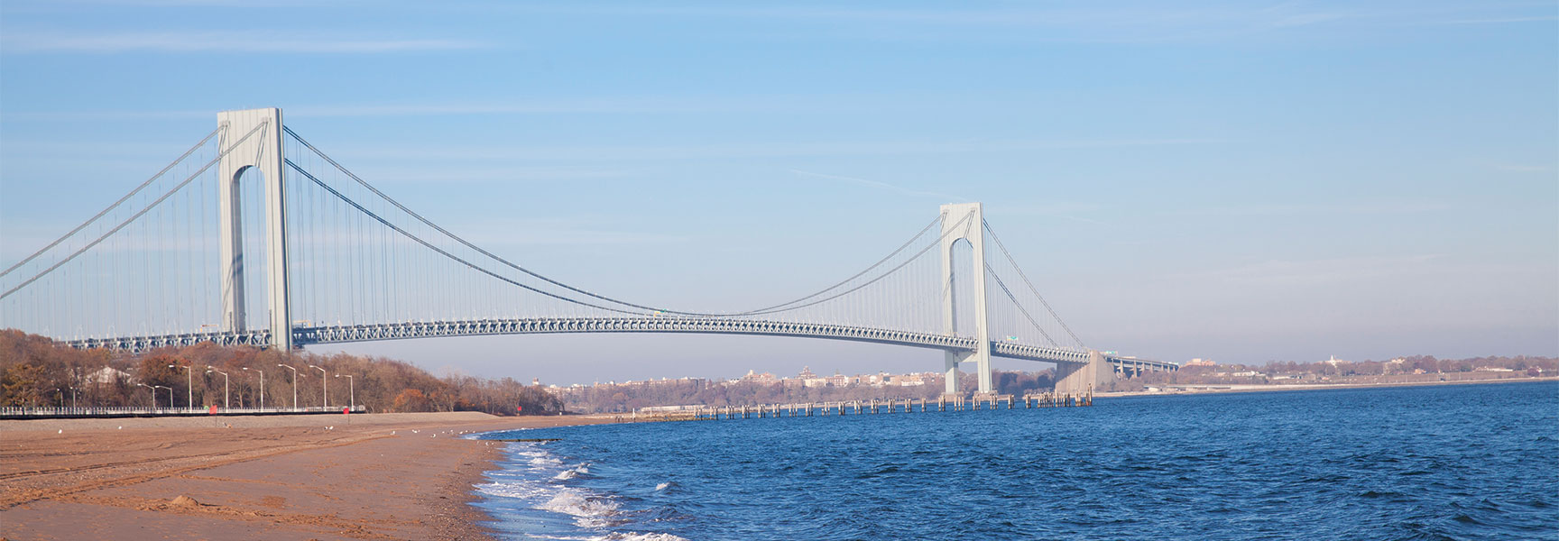 A view of the Verrazzano-Narrows Bridge from a sandy New York City beach, capturing the surf and shoreline under a clear sky.