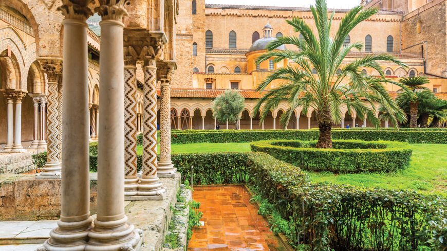 Historic courtyard garden in Sicily, Italy, featuring ornate stone columns and lush palm trees, showcasing the region's rich architectural and cultural history.