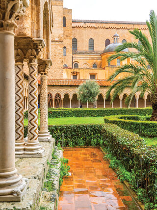 Historic courtyard garden in Sicily, Italy, featuring ornate stone columns and lush palm trees, showcasing the region's rich architectural and cultural history.