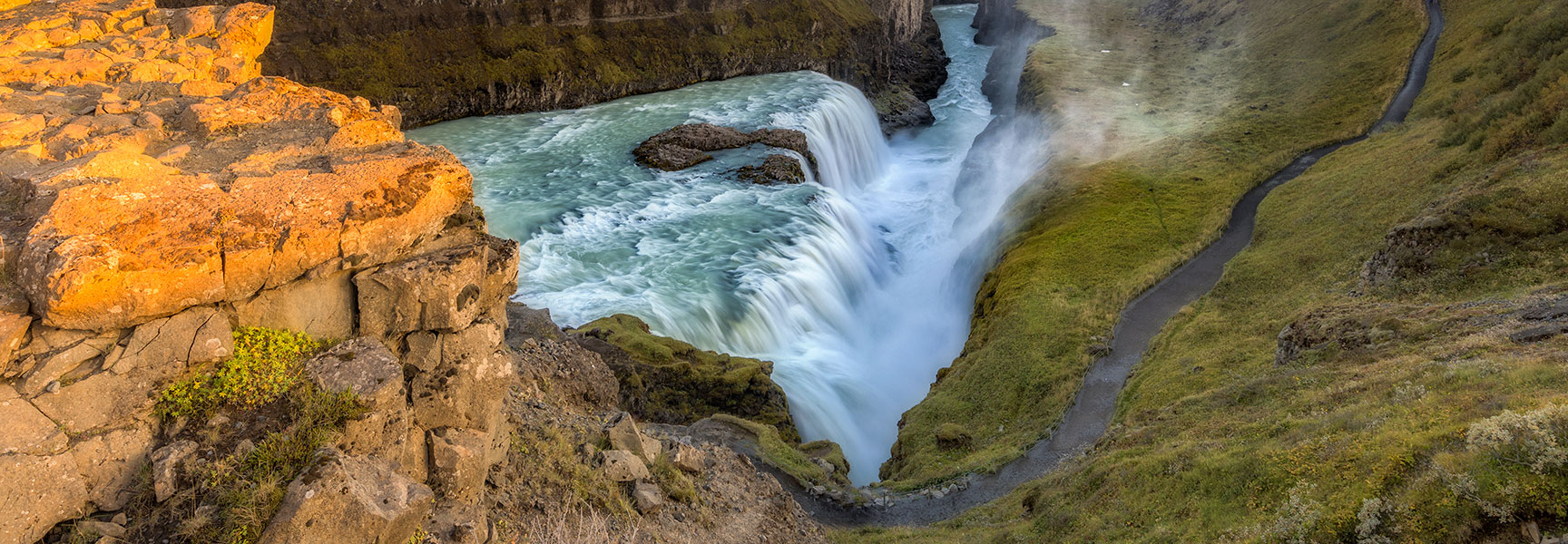 A wide view of the magnificent Gullfoss Falls in Iceland, cascading into a canyon with a trail winding along the grassy cliffs.