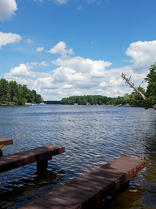 A wooden dock on a lake in Wisconsin, with a tree-lined shore in the distance under a blue sky with clouds.