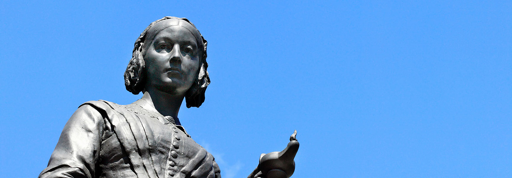 A bronze statue of Florence Nightingale holding a lamp, viewed from below against a clear, bright blue sky.