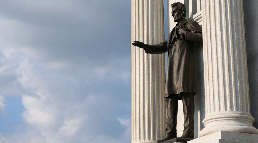 A bronze statue of Abraham Lincoln stands next to a large fluted column, gesturing with his hand under a cloudy sky.