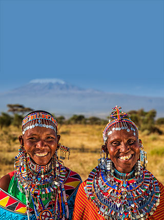 Two Maasai women in traditional colorful beaded jewelry smile in a field in Kenya, with a mountain in the background.