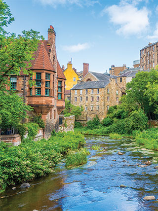 Historic stone buildings of Dean Village in Edinburgh, Scotland, line the lush green banks of the Water of Leith under a blue sky.