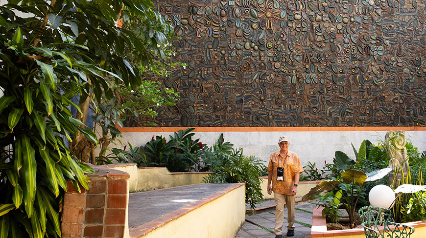 A man walks through a lush courtyard in Havana, Cuba, in front of a large, textured wall mural.