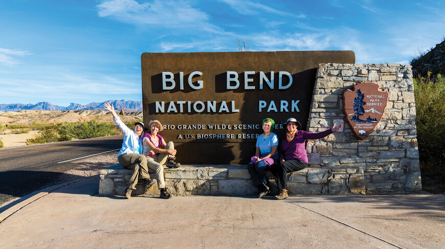 Four women pose and wave in front of the Big Bend National Park entrance sign in the rugged landscape of Texas.