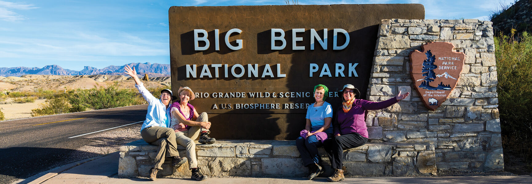 Four women pose and wave in front of the Big Bend National Park entrance sign in the rugged landscape of Texas.