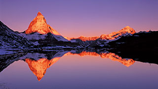 The iconic Matterhorn mountain in Switzerland is illuminated by sunrise, its peak reflected in the calm waters of a lake below.