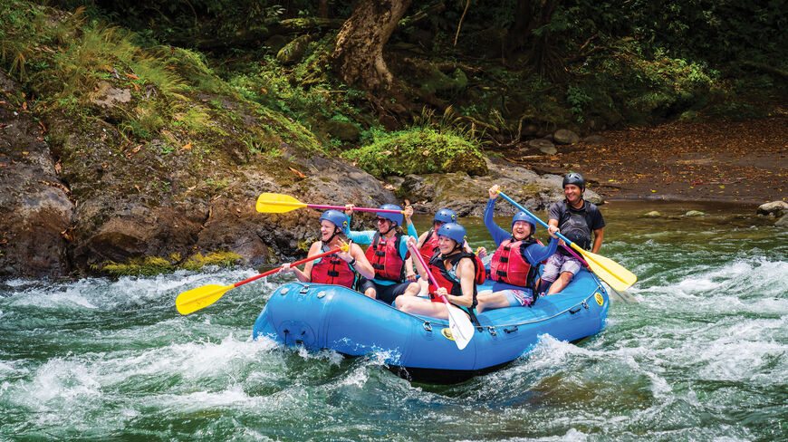 Women on a multi-sport adventure in Costa Rica navigate white water rapids in a blue raft through a tropical forest.