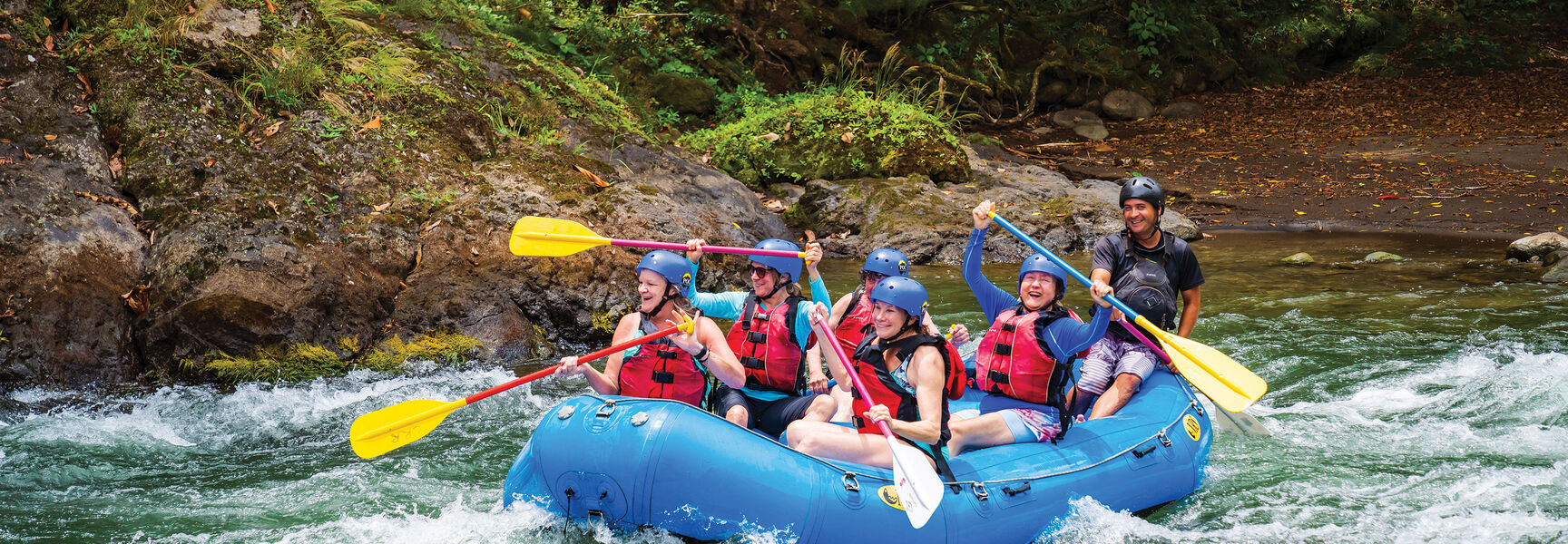 Women on a multi-sport adventure in Costa Rica navigate white water rapids in a blue raft through a tropical forest.
