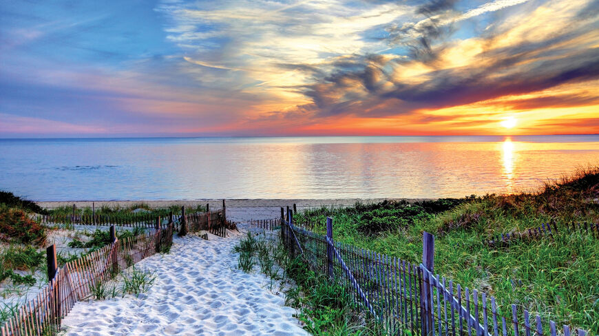 A sandy path lined with wooden fences leads to a beach in Cape Cod, Massachusetts, under a vibrant sunset sky.