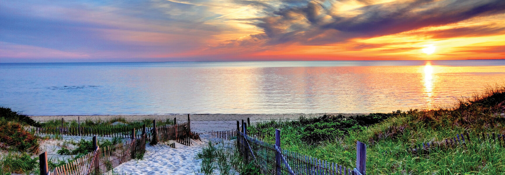 A sandy path lined with wooden fences leads to a beach in Cape Cod, Massachusetts, under a vibrant sunset sky.
