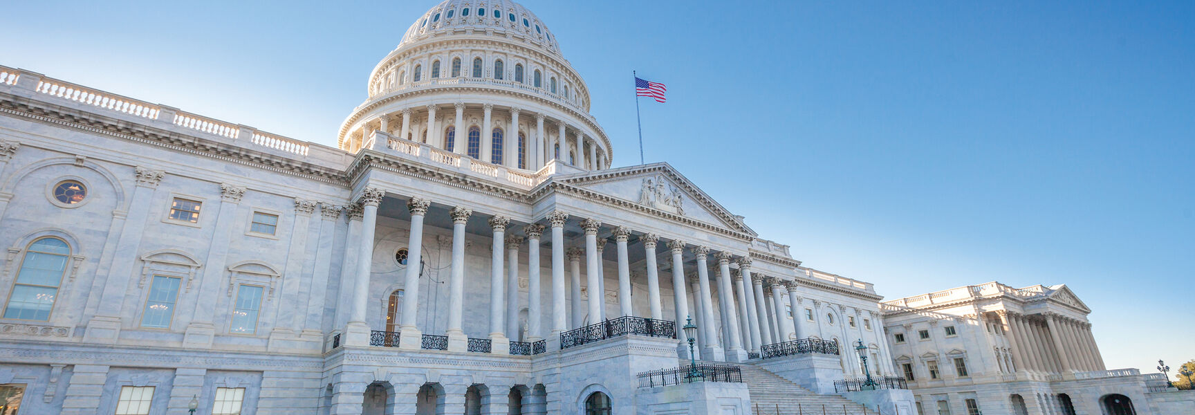 A low-angle, historic view of the United States Capitol building in Washington, D.C., featuring its white dome and marble columns under a clear sky.