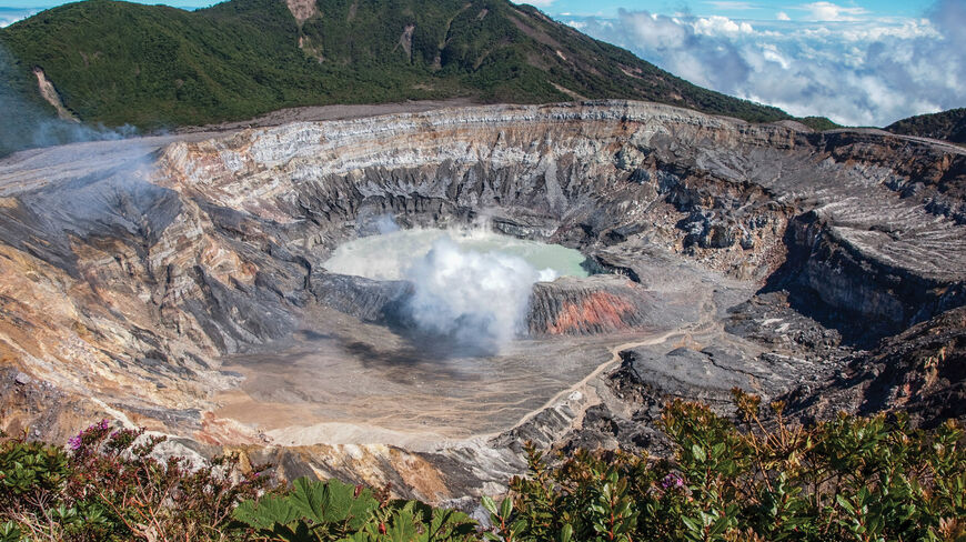 A massive volcanic crater in Costa Rica featuring a steaming turquoise lake at its center, surrounded by steep rocky walls and lush green mountains.