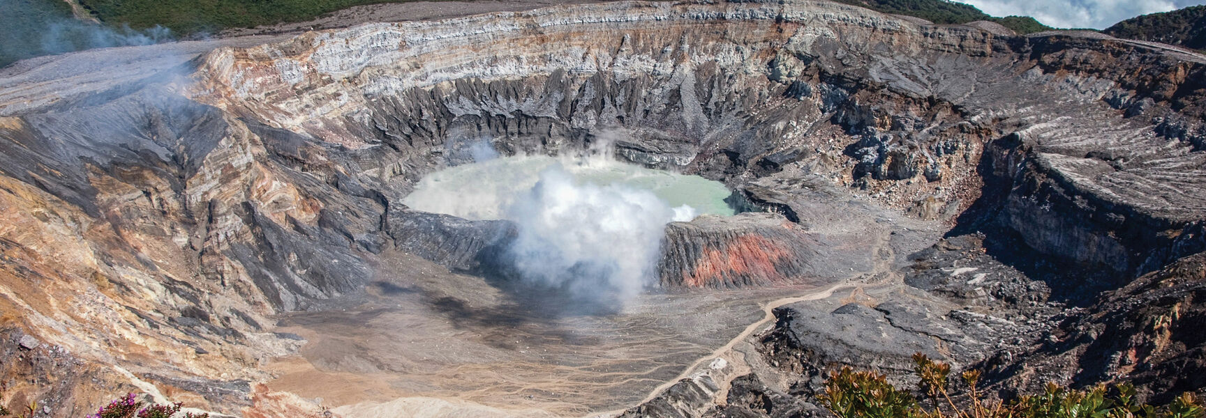 A massive volcanic crater in Costa Rica featuring a steaming turquoise lake at its center, surrounded by steep rocky walls and lush green mountains.
