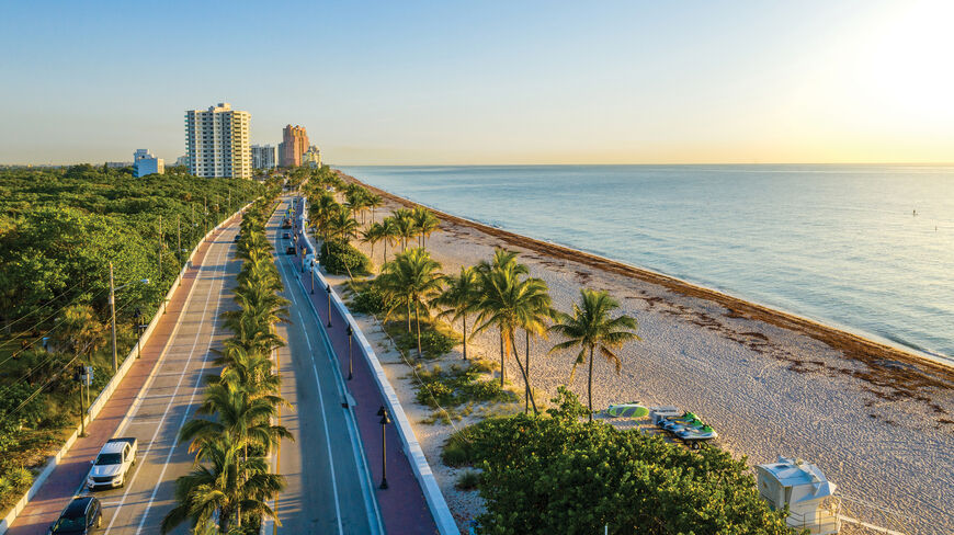 An aerial view of a palm-lined coastal road in Miami, Florida, running alongside a sandy beach and the calm blue ocean at sunset.