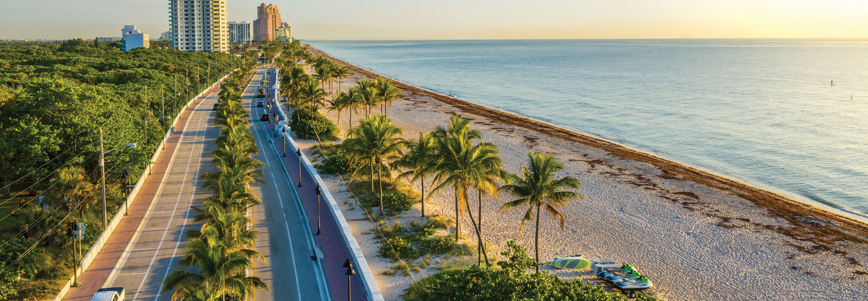An aerial view of a palm-lined coastal road in Miami, Florida, running alongside a sandy beach and the calm blue ocean at sunset.